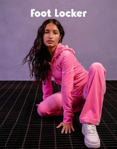 Brunette woman wearing a pink velour tracksuit and white sneakers poses on a metal grate against a lilac background. Foot Locker logo.