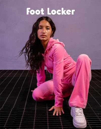 Brunette woman wearing a pink velour tracksuit and white sneakers poses on a metal grate against a lilac background. Foot Locker logo.