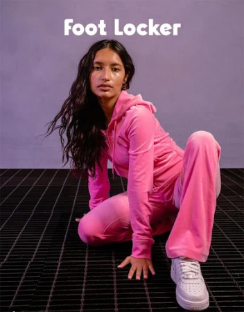Brunette woman wearing a pink velour tracksuit and white sneakers poses on a metal grate against a lilac background. Foot Locker logo.