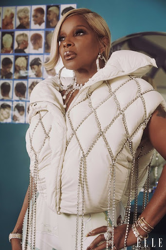 Mary J. Blige poses in a cream-colored puffy vest adorned with rhinestones, with a hair style chart visible in the background.