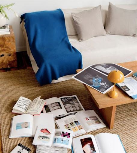 Bright indoor living room with sofa, wooden coffee table, throw blanket, lamp, and multiple open books on a jute rug.