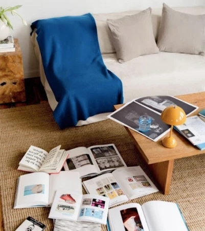 Bright indoor living room with sofa, wooden coffee table, throw blanket, lamp, and multiple open books on a jute rug.