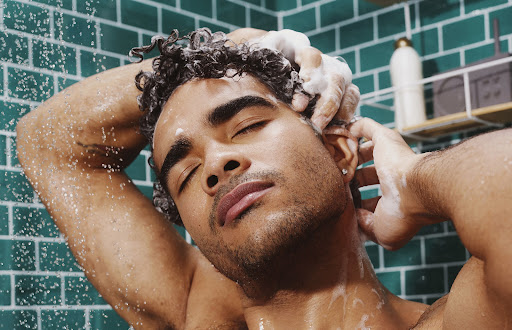 Close-up of a shirtless adult male washing his curly hair in a tiled shower with closed eyes, creating a serene and intimate grooming scene.