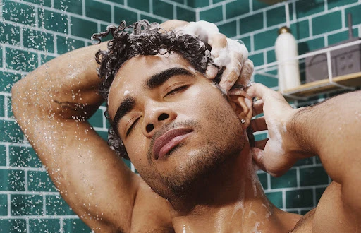 Close-up of a shirtless adult male washing his curly hair in a tiled shower with closed eyes, creating a serene and intimate grooming scene.