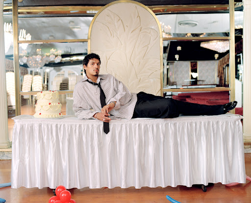 An adult male with dark hair is laying on a white, ruffled tablecloth-covered table next to a white cake with red decoration.