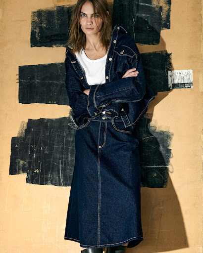 Model in dark denim jacket and matching skirt standing against a textured wall with painted black brushstrokes