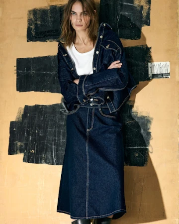 Model in dark denim jacket and matching skirt standing against a textured wall with painted black brushstrokes