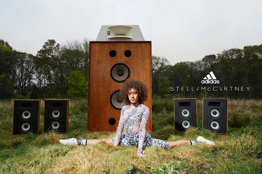 Athletic woman in patterned bodysuit does the splits in a field with large speakers, promoting Adidas Stella McCartney collection.