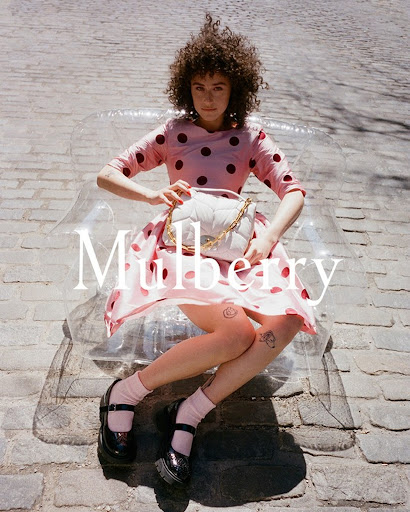 Brunette woman in pink polka dot dress and black patent shoes poses with white purse on a cobblestone city street.