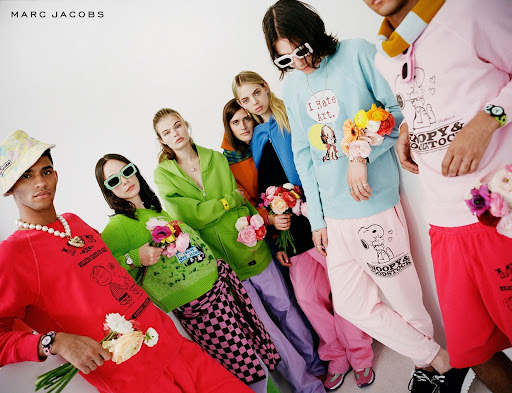 Group of six fashion models wearing colorful casual outfits with flower bouquets. Marc Jacobs brand visible. Bright studio lighting.
