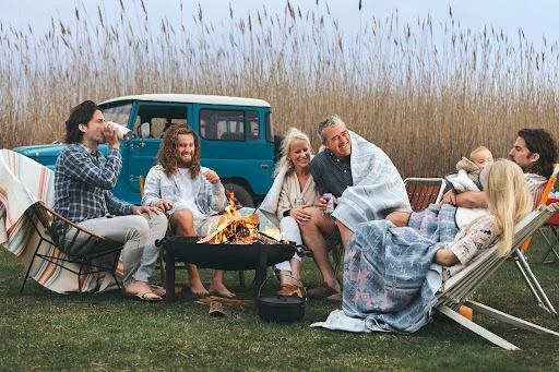Group of adults and children gather around a campfire on a grassy field, with a blue SUV and tall grass in the background.