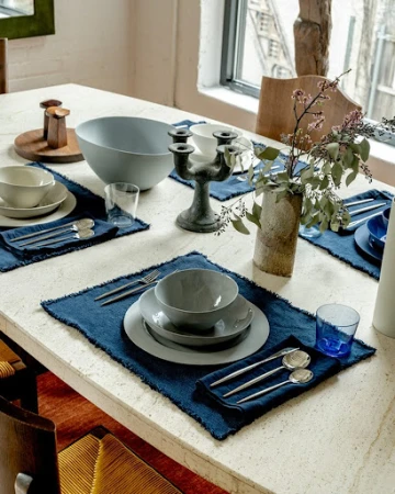 Styled dining table setting with dark blue placemats, grey ceramic bowls, and a ceramic vase with branches.