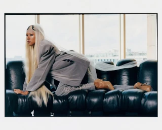 Model with long blonde hair and sheer veil posing on a black leather sofa wearing an oversized grey suit and beige heels.