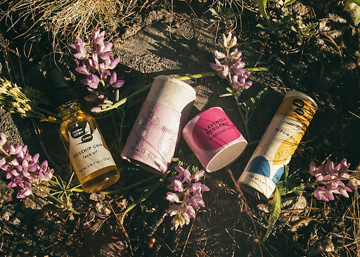 Four tubes and bottles of natural skincare products, featuring pastel labels and flower blossoms, are displayed outdoors on the ground.