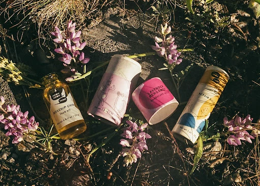 Four tubes and bottles of natural skincare products, featuring pastel labels and flower blossoms, are displayed outdoors on the ground.
