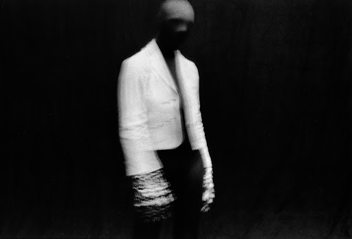Abstract fashion portrait of a blurred figure in a white blazer and skirt against a dark background, artistic monochrome aesthetic.