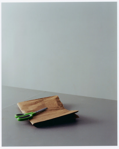 Still life photo of a brown paper bag and green scissors on a grey surface against a plain grey background, simple composition.