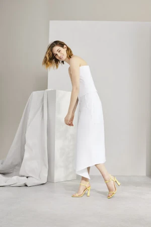 A smiling woman wearing a white strapless top and skirt with yellow heeled sandals in a studio setting by Kate Stein
