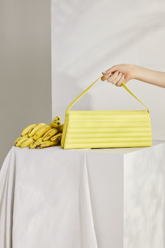 A person's arm holding a bright yellow handbag with a handle next to a bunch of ripe yellow bananas on a draped white surface.