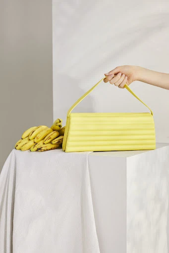 A person's arm holding a bright yellow handbag with a handle next to a bunch of ripe yellow bananas on a draped white surface.