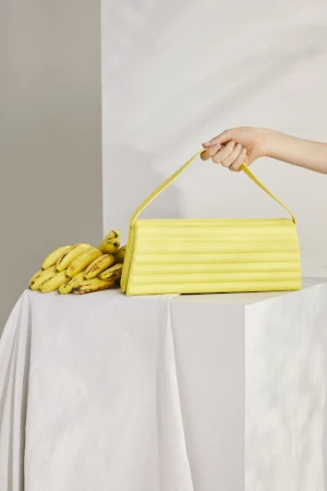 A person's arm holding a bright yellow handbag with a handle next to a bunch of ripe yellow bananas on a draped white surface.