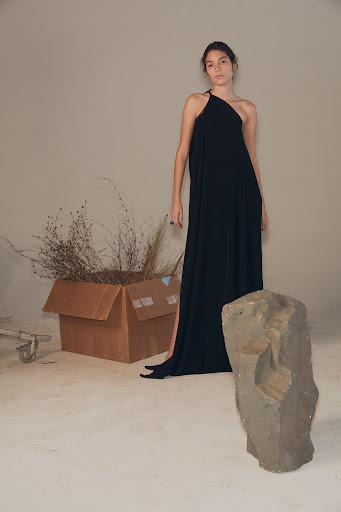 A woman in a black dress stands next to a cardboard box filled with dry grasses and a rough stone sculpture in a studio setting.