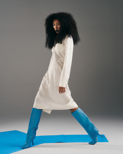 Fashionable woman with dark curly hair posing in a long sleeve white dress with blue knee-high boots against a gray background.