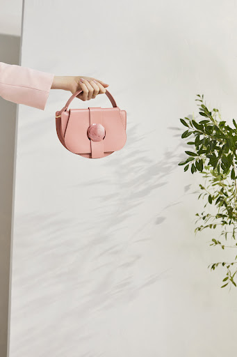 Hand holds a pink purse against a white wall with plant shadows. Close-up fashion studio shot. Great for beauty and style search.