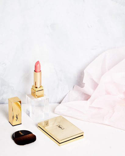 Elegant beauty still life featuring YSL lipstick, powder compact, and brush on a white surface with a soft pink and white backdrop.
