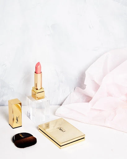 Elegant beauty still life featuring YSL lipstick, powder compact, and brush on a white surface with a soft pink and white backdrop.