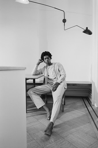 A young Black man seated barefoot on a wooden chair in an interior room with a wall-mounted adjustable lamp and parquet floor.