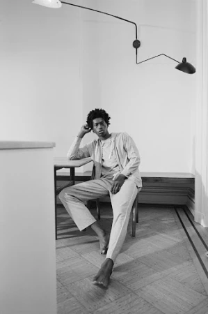 A young Black man seated barefoot on a wooden chair in an interior room with a wall-mounted adjustable lamp and parquet floor.