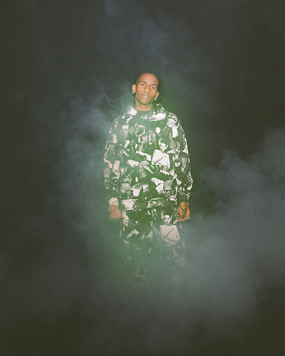 Adult male stands in a photo studio, illuminated by soft lighting and surrounded by smoke, wearing a patterned hoodie and pants.