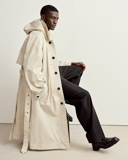 An adult male sits, modeling a long, hooded trench coat in an indoor setting with a plain, light-colored wall behind him, showcasing fashion apparel.
