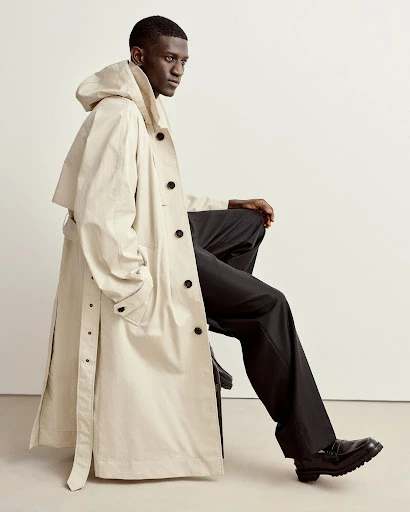 An adult male sits, modeling a long, hooded trench coat in an indoor setting with a plain, light-colored wall behind him, showcasing fashion apparel.