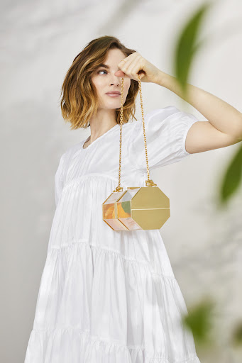A woman in a white dress holds an octagonal handbag with a gold chain, posing in an indoor setting with leaf shadows.