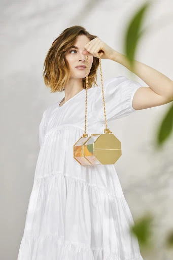 A woman in a white dress holds an octagonal handbag with a gold chain, posing in an indoor setting with leaf shadows.