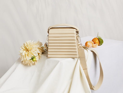Still life of a cream-colored purse with geometric details, arranged with white dahlias and vibrant tangerines on a white tablecloth.