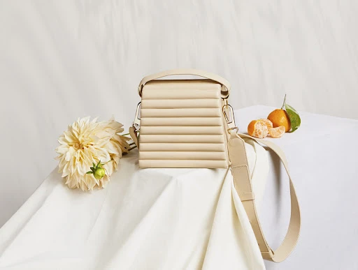 Still life of a cream-colored purse with geometric details, arranged with white dahlias and vibrant tangerines on a white tablecloth.