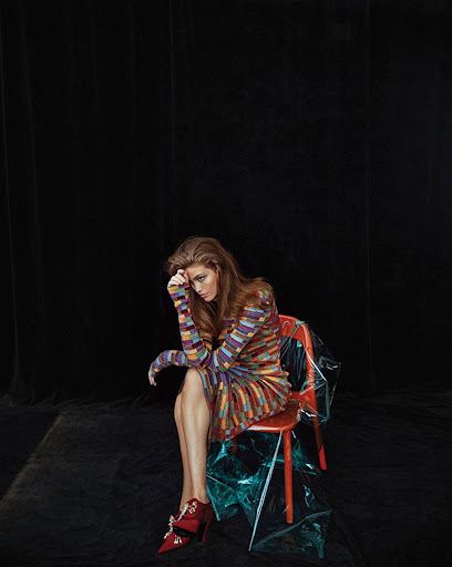 Model with long reddish-brown hair wearing a multicolored patterned dress and red shoes seated on a red chair against black backdrop