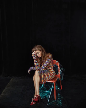 Model with long reddish-brown hair wearing a multicolored patterned dress and red shoes seated on a red chair against black backdrop