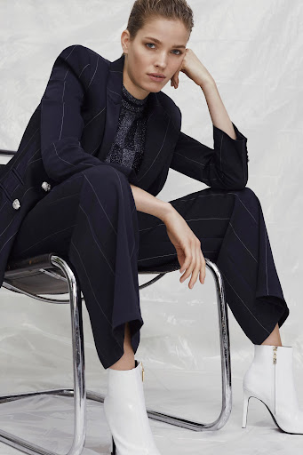 Woman in a navy pin-striped suit sits on a chrome chair wearing white ankle boots in a studio setting with a crinkled white background.