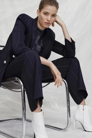 Woman in a navy pin-striped suit sits on a chrome chair wearing white ankle boots in a studio setting with a crinkled white background.