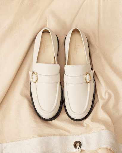 Studio shot of a pair of white leather penny loafers with gold buckles on a wrinkled tan fabric backdrop, fashion footwear product shot.