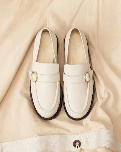 Studio shot of a pair of white leather penny loafers with gold buckles on a wrinkled tan fabric backdrop, fashion footwear product shot.
