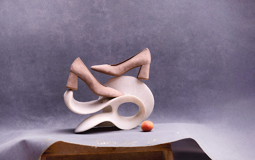 Elegant beige high-heeled shoes gracefully balanced on a white sculptural stand, complemented by a solitary peach, against grey backdrop.