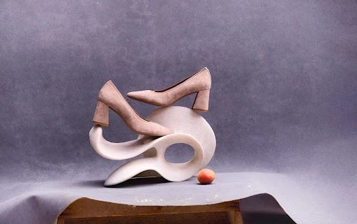 Elegant beige high-heeled shoes gracefully balanced on a white sculptural stand, complemented by a solitary peach, against grey backdrop.
