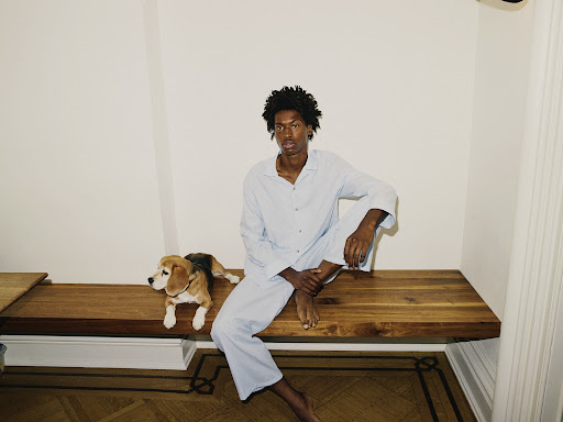 An adult male in pajamas sits on a wooden bench with a beagle dog in an indoor setting, illuminated by bright, soft lighting.