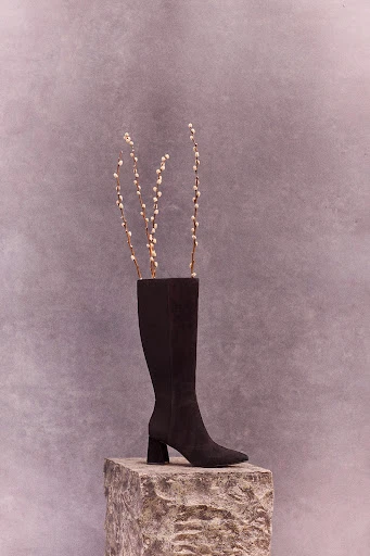 Knee-high black suede boot on a stone block with pussy willow branches inside, against a textured grey background, vertical.