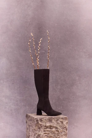Knee-high black suede boot on a stone block with pussy willow branches inside, against a textured grey background, vertical.